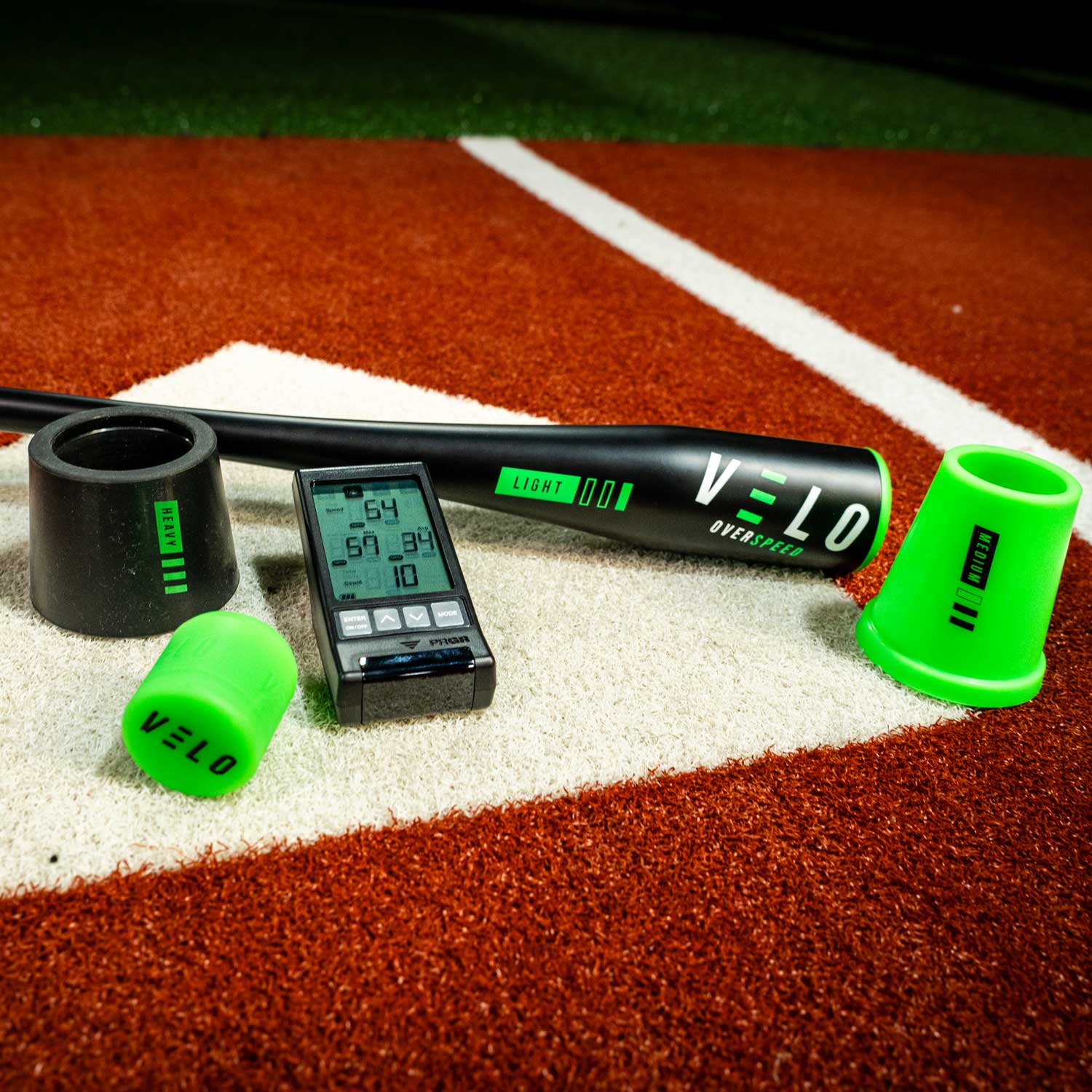 A set of VELO Overspeed baseball training equipment including a black and green training bat with green and white accents, a PRGR digital bat speed radar monitor, with medium green weight and heavy black weight, placed on a red artificial turf with white baseball diamond markings
